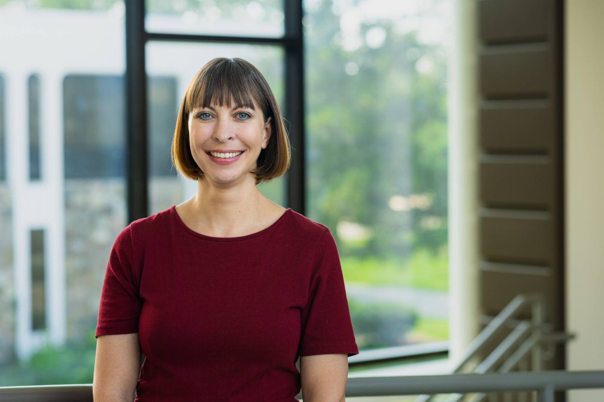 A smiling woman with short hair and bangs, wearing a burgundy top, standing in front of large windows with a blurred green outdoor background.